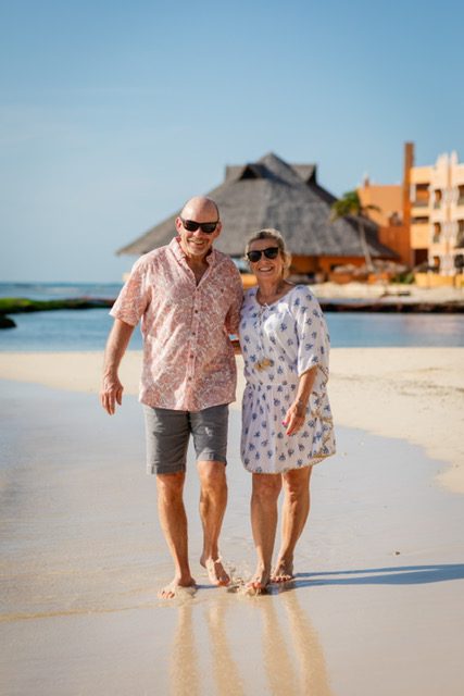 A couple walking hand in hand on a beach with buildings in the background.