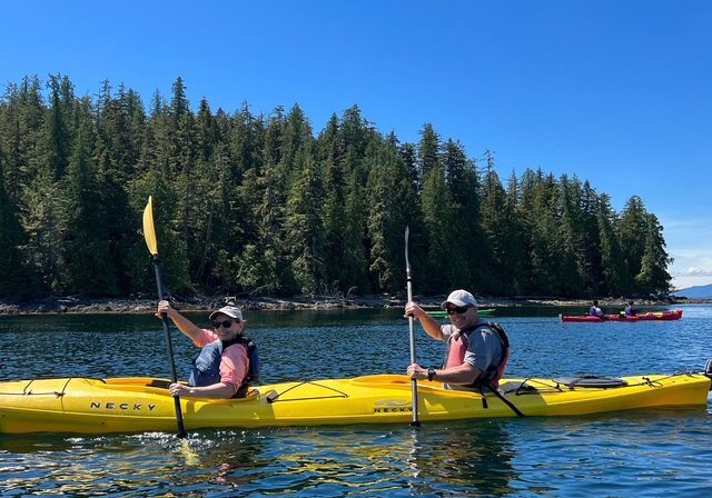 Two people kayaking on a calm lake near a forested shore under clear blue skies.