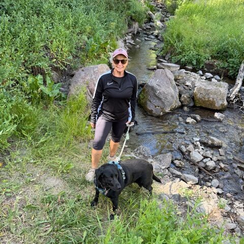 Person with sunglasses and dog near a stream in a green natural setting.